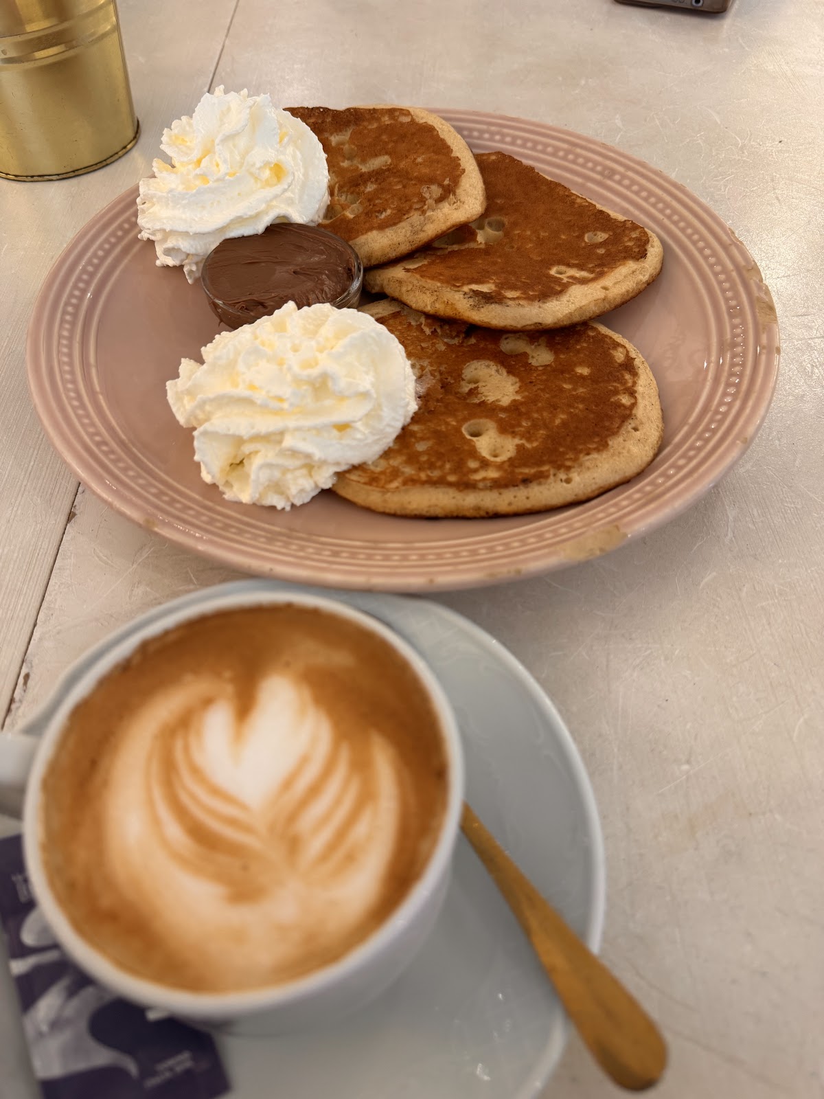Panqueques y café. Se presenta un plato con tres panqueques, acompañados de crema batida y una porción de chocolate, junto a una taza de café.